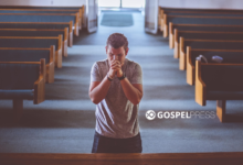 Homem ajoelhado, de costas, orando sozinho em um banco de igreja histórica, com vitrais coloridos iluminando o ambiente.