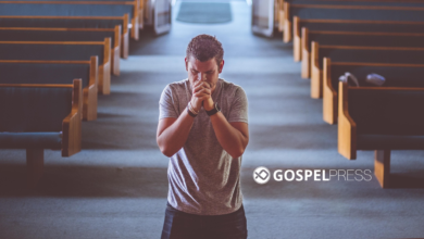 Homem ajoelhado, de costas, orando sozinho em um banco de igreja histórica, com vitrais coloridos iluminando o ambiente.