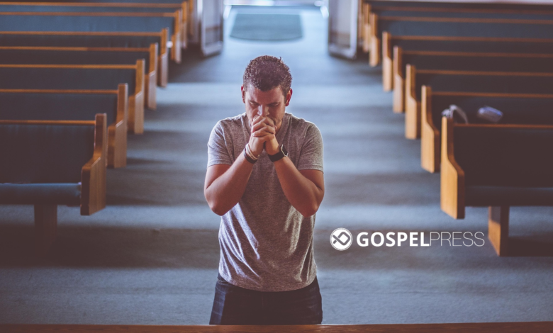 Homem ajoelhado, de costas, orando sozinho em um banco de igreja histórica, com vitrais coloridos iluminando o ambiente.